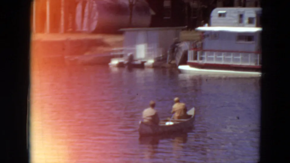 1969 Minnesota river view with two men in a canoe, a docked houseboat, and a small structure along the shore.