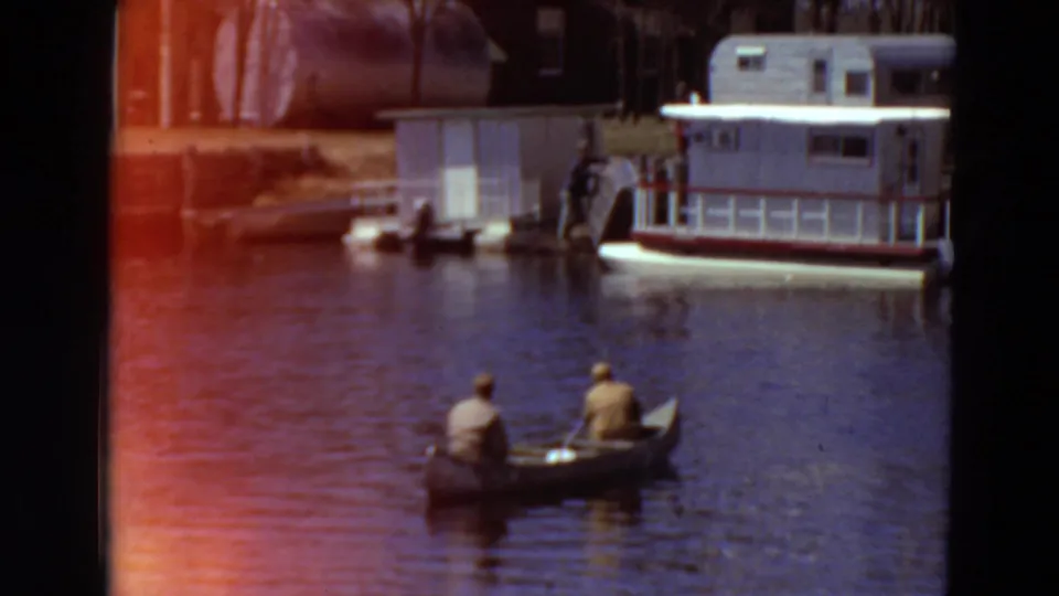 1969 Minnesota 8mm frame of two men in a canoe below a docked houseboat and small shoreline building.