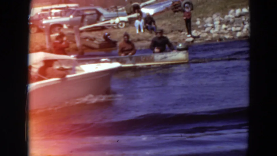 1969 Minnesota river scene with a motorboat passing people gathered beside cars and boats along a rocky shoreline.