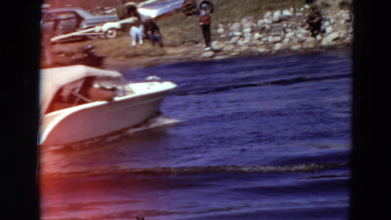 A 1969 Minnesota 8mm frame shows a white motorboat moving past a rocky riverbank with bystanders and older cars above the water.