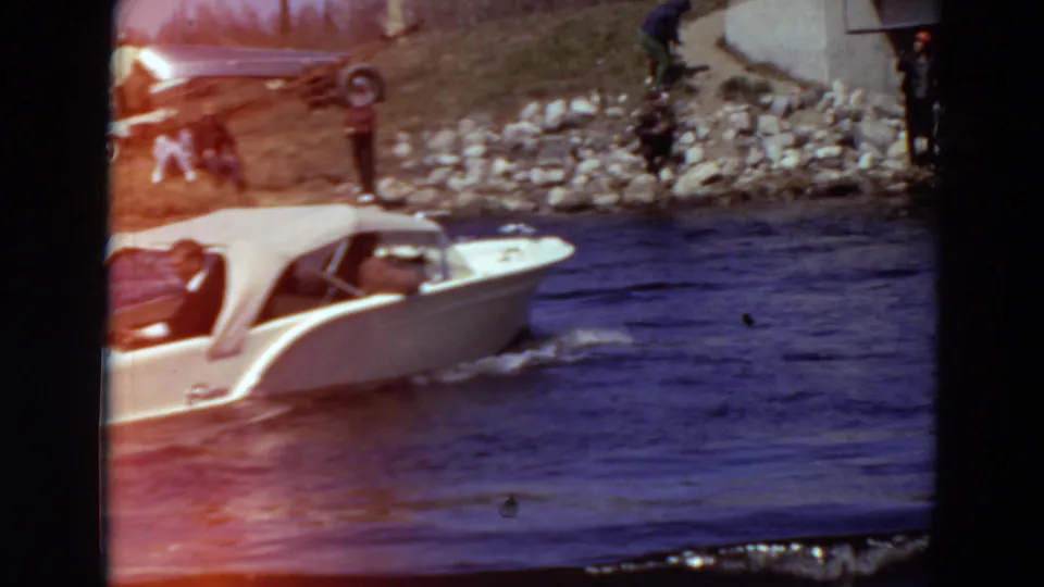 The white runabout continues along the Minnesota river, with people watching from a rocky bank and older cars parked above.