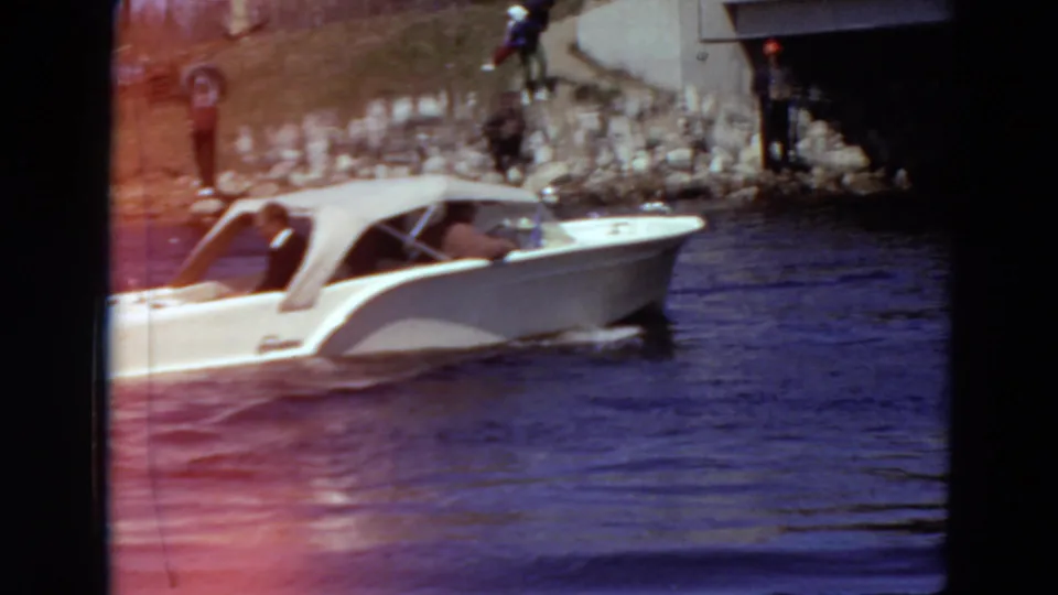 A 1969 home-movie frame shows the motorboat nearing the bridge area while distant spectators stand along the rocky shoreline.