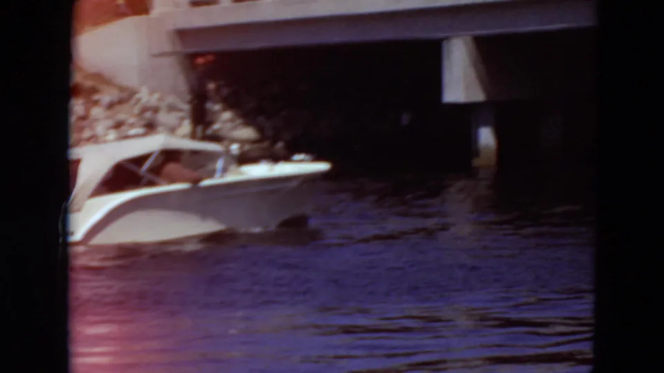 The white motorboat enters the shadow of a concrete bridge, with the river surface and rocky bank still partly visible in the 8mm frame.