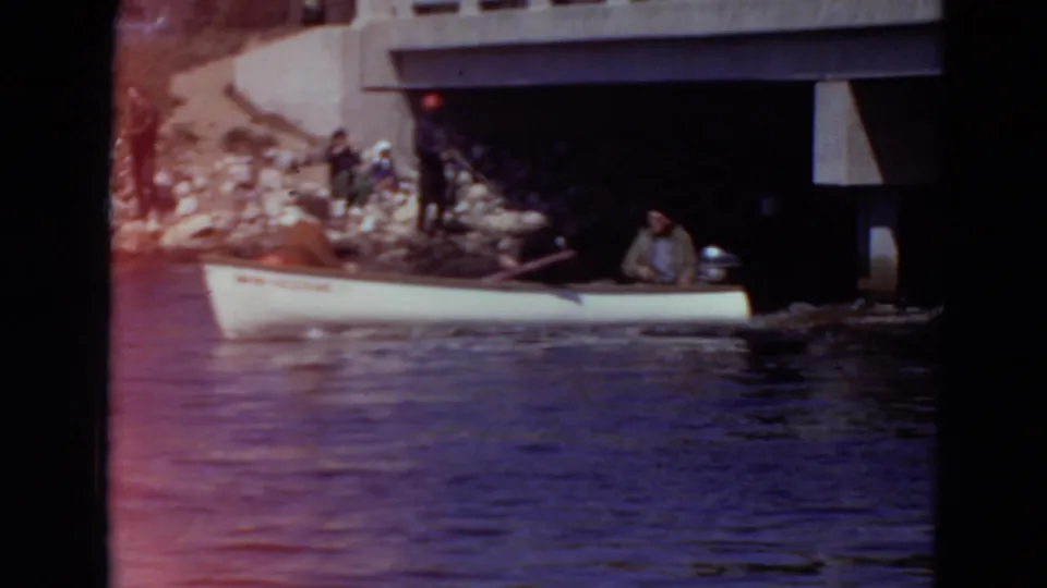 1969 Minnesota home-movie frame of a canoe with people paddling near rocks and bridge supports along the water.