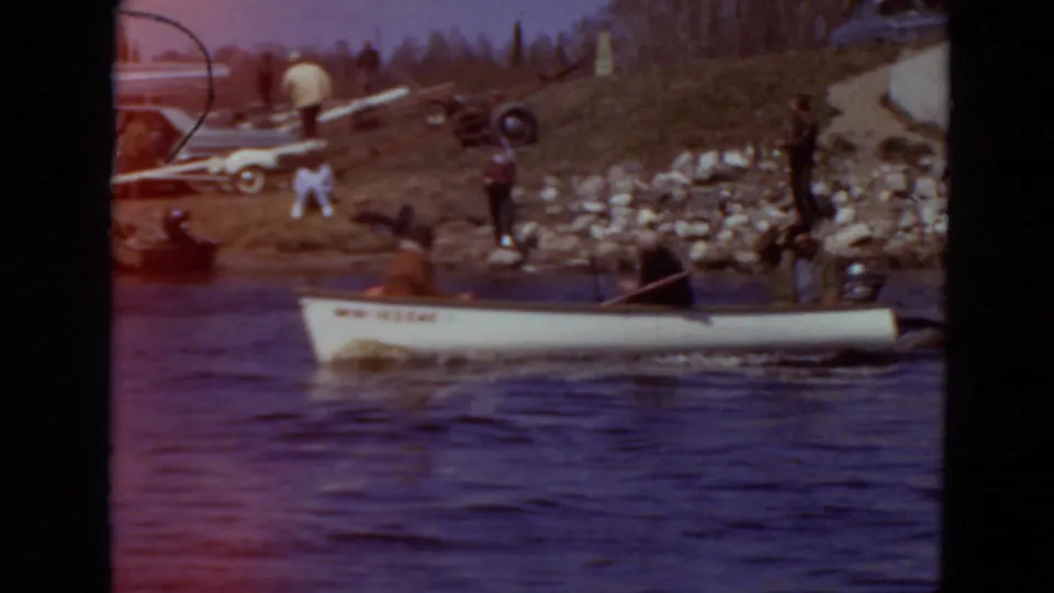 1969 Minnesota frame showing a canoe on rippled water, shoreline figures, rocks, and part of another boat at left.