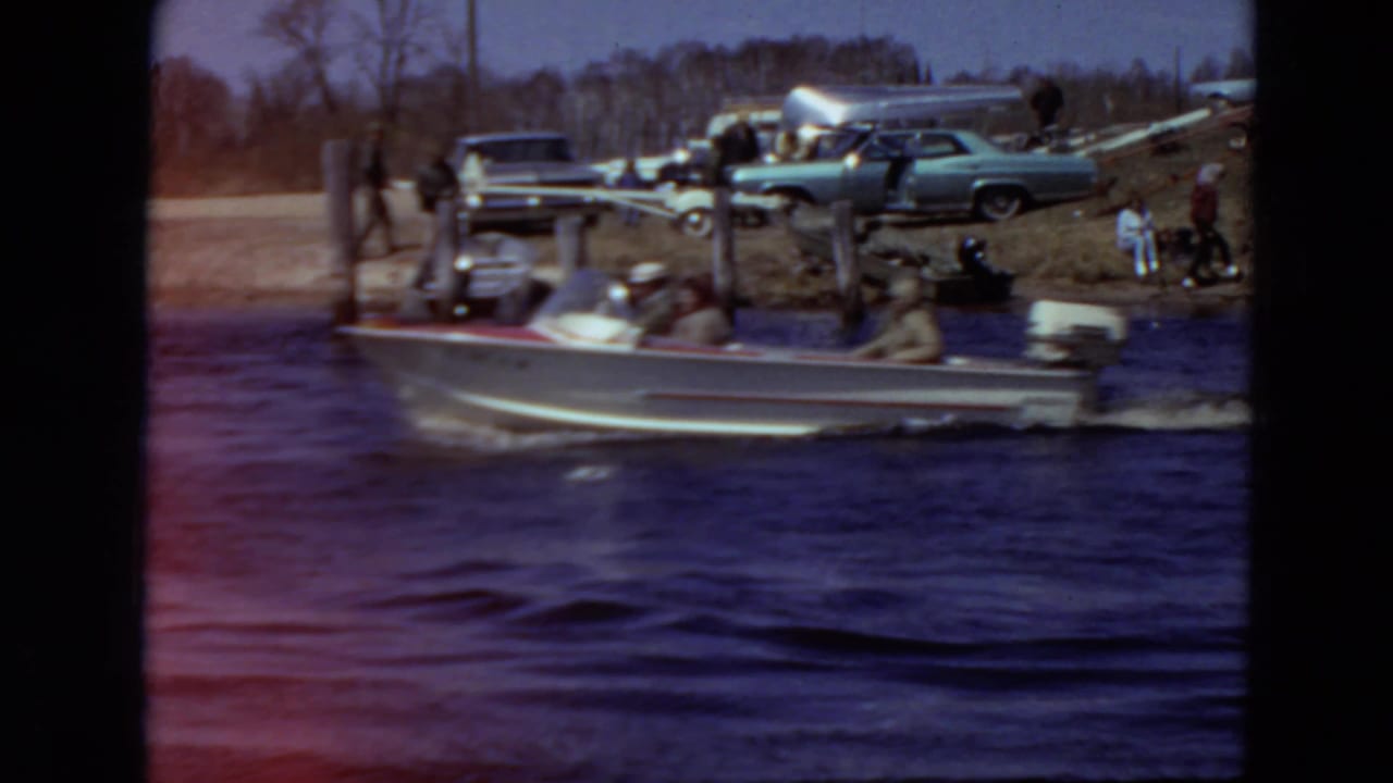 Small motorboat passes near a riverbank with parked cars and people on shore.