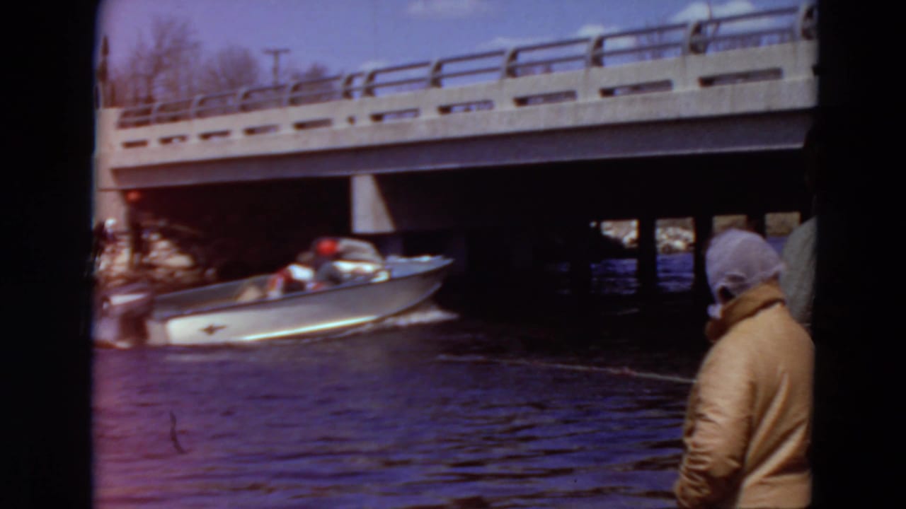 Motorboat speeds toward a concrete bridge while a person in a hooded coat watches from shore.