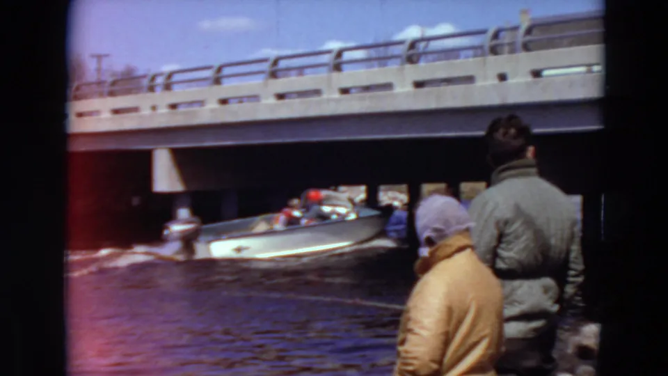 1969 Minnesota home-movie frame showing a small motorboat under a bridge, viewed past two people standing on the riverbank.