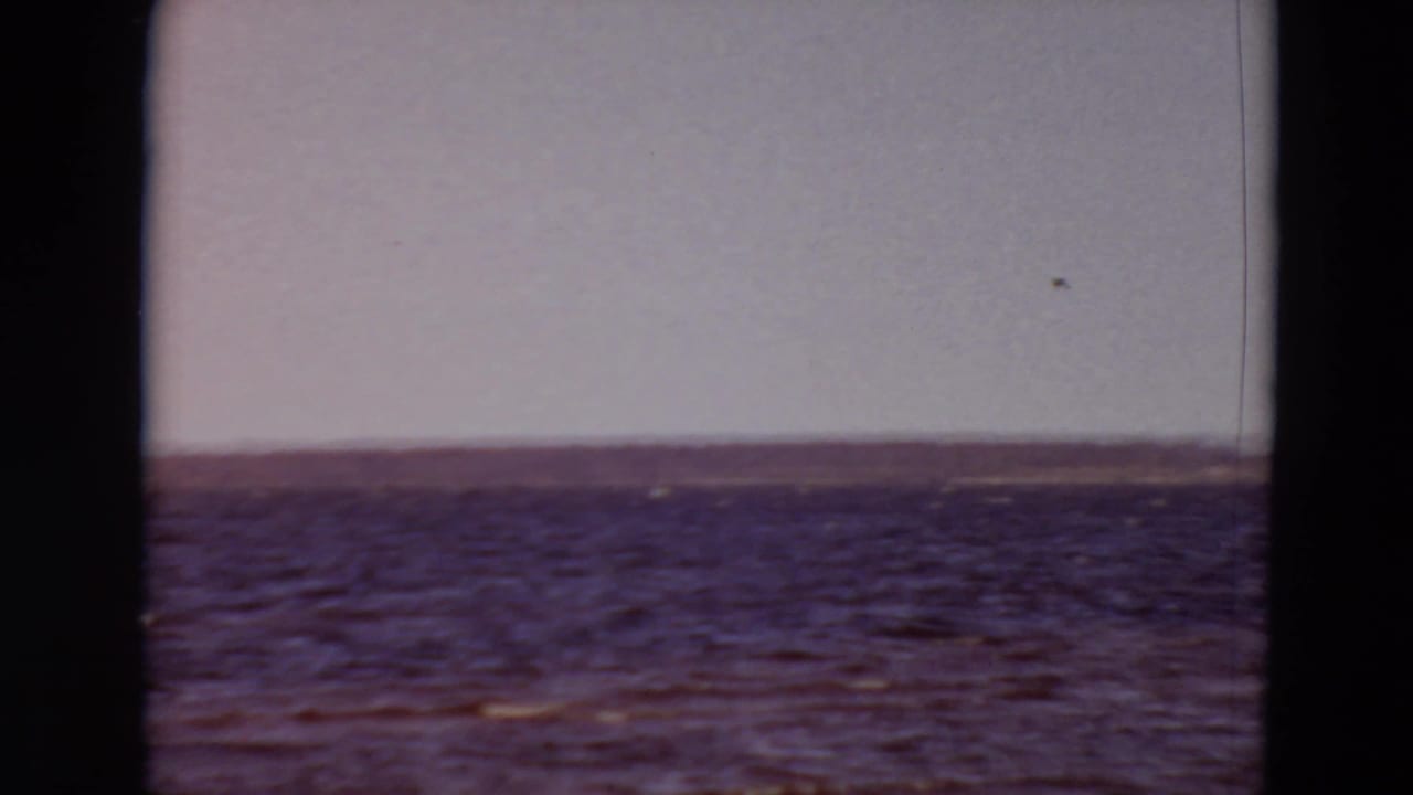1969 8mm home-movie frame of rippled water and a distant shoreline, seen through dark film gate edges.