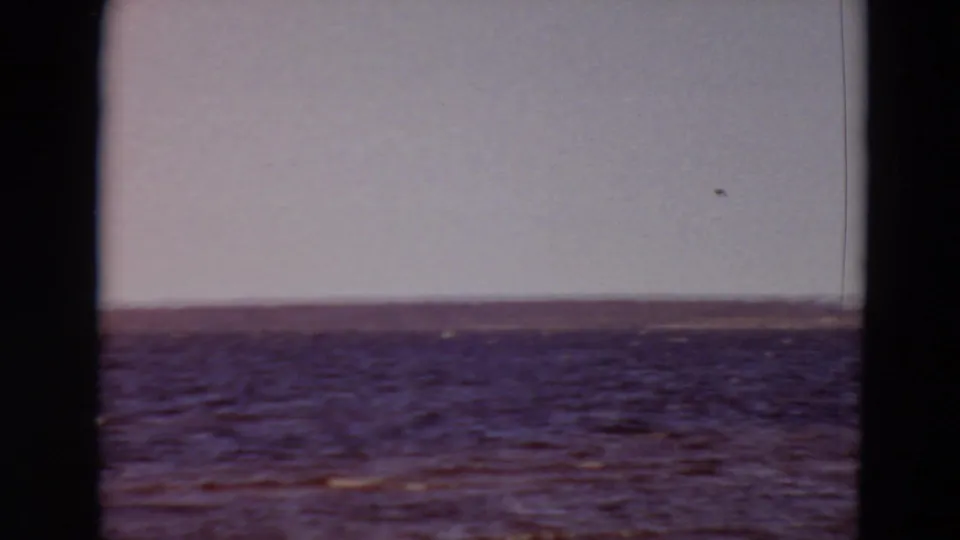 1969 8mm home-movie frame of rippled water and a distant shoreline, seen through dark film gate edges.