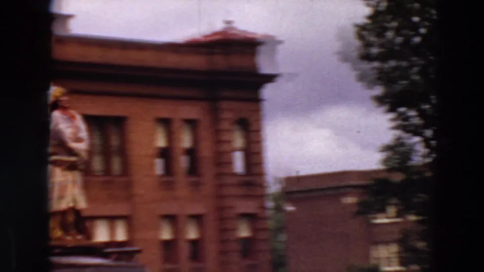 Minnesota 1969 8mm frame showing a brick building corner, rooftop statue, nearby tree, and soft overcast sky.