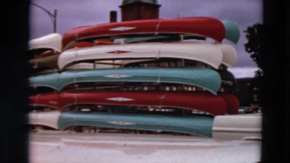 1969 Minnesota home-movie frame of red, white, teal, and green canoes stacked on racks under an overcast sky.