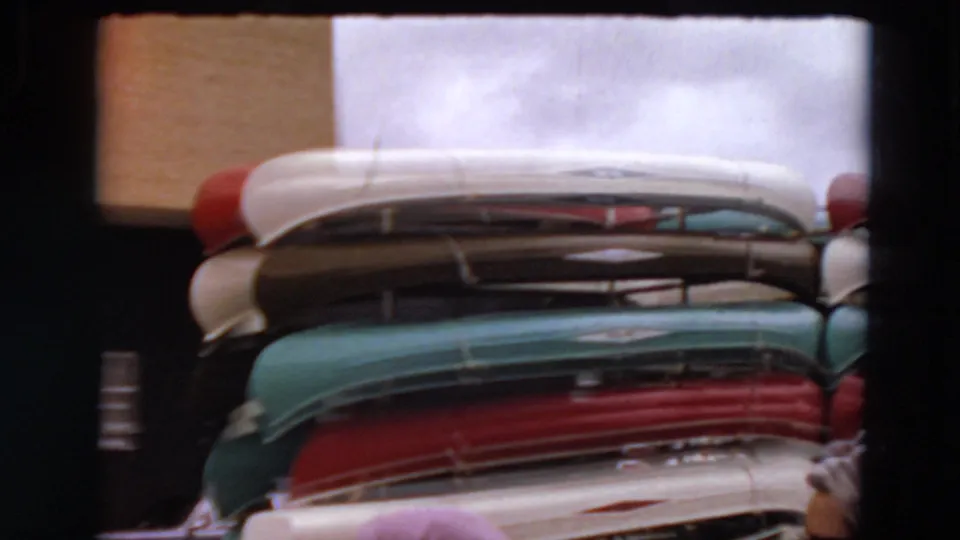 Home-movie frame of colorful canoes stacked near a building, with partial human figures visible at the lower and right edges.