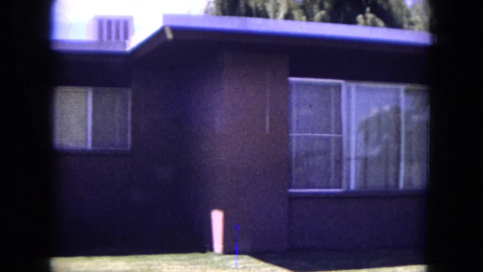 1970 White Sands, Florida frame of the same suburban house exterior, with shaded siding, bright lawn, and large curtained windows.