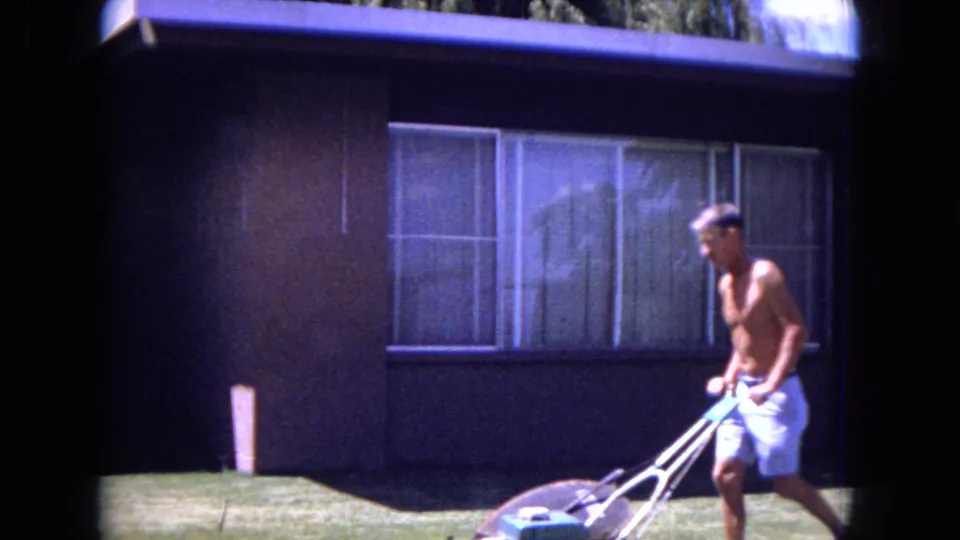 1970 White Sands, Florida frame showing an adult man mowing the lawn with a push mower in front of a suburban house.