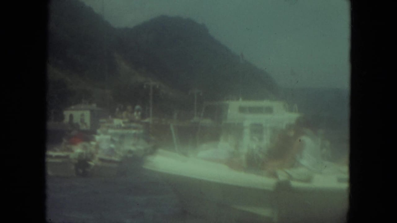 People in a small white boat near a docked vessel with hills behind them.