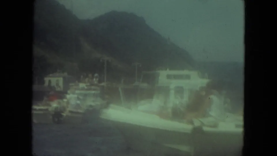 1971 Mississippi-labeled home-movie frame of people aboard a small white boat near a docked vessel and wooded hills.