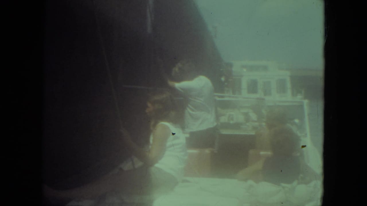 1971 Mississippi River frame with a man in a white shirt standing near rigging while passengers sit in a small boat beside open water.