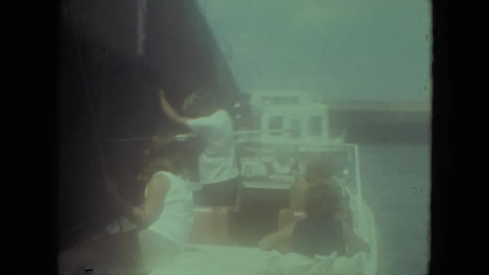 1971 Mississippi River home-movie frame showing seated passengers and a standing man working near a dark sail or rope on a small boat.