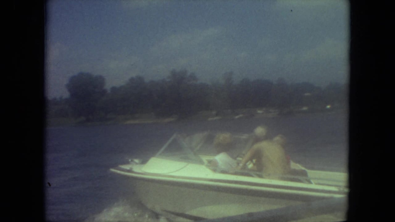 Person seated in a small white motorboat on a lake with shoreline trees in the distance.