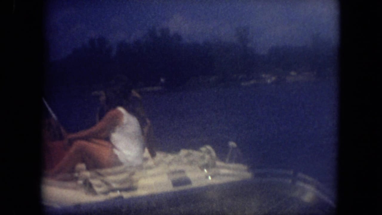 Dusk-toned 1971 Mississippi frame with passengers relaxing in a small motorboat on a lake, shoreline trees visible beyond.