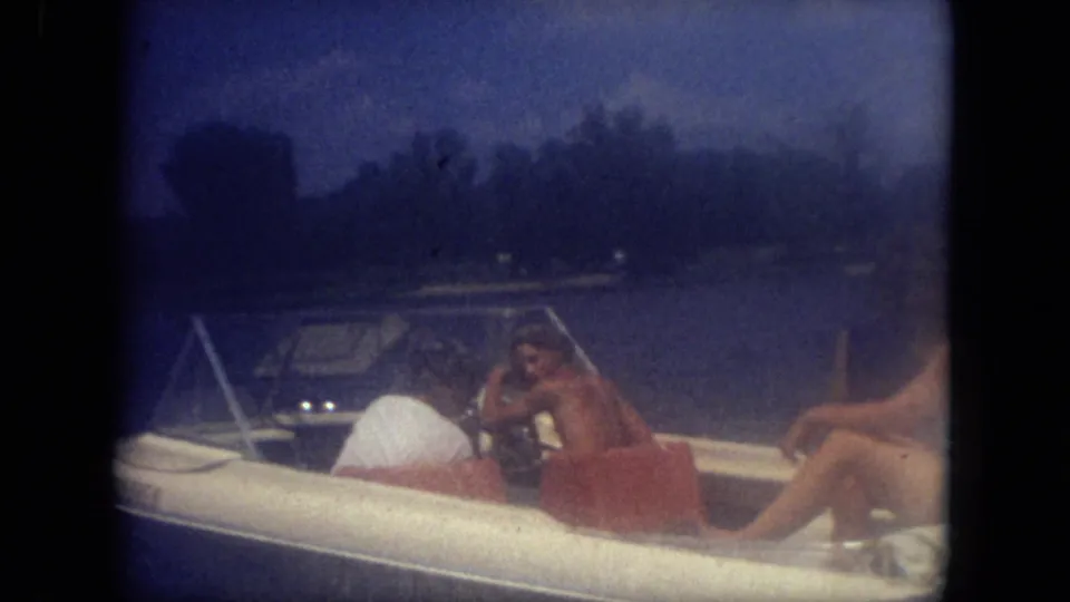 1971 Mississippi home-movie frame showing three adults in swimwear aboard a white motorboat with lake water and shoreline behind them.