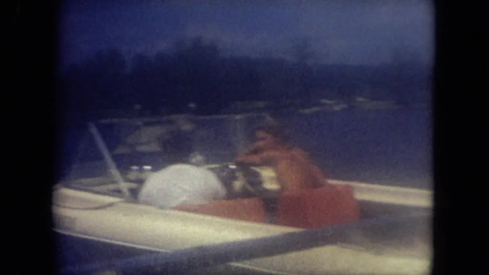 Near-dusk 1971 Mississippi frame with adults aboard a small white motorboat on lake water, seen through soft, blue-toned 8mm exposure.