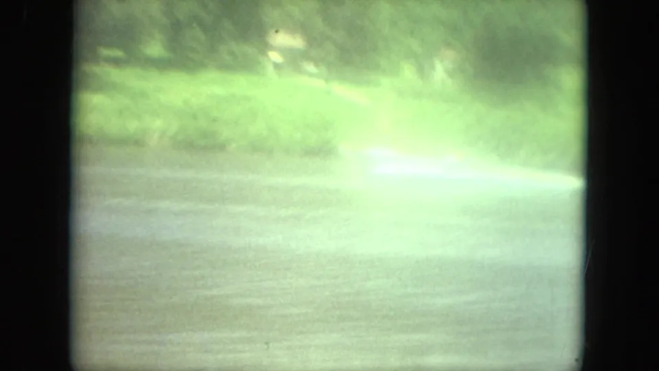 Distant 1971 Mississippi lake view of a small boat moving fast across water, with bright wake and green shoreline.