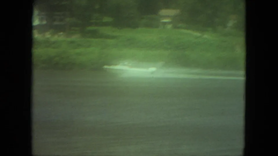 Wide 1971 Mississippi 8mm frame of a distant boat cutting across lake water near a green bank.