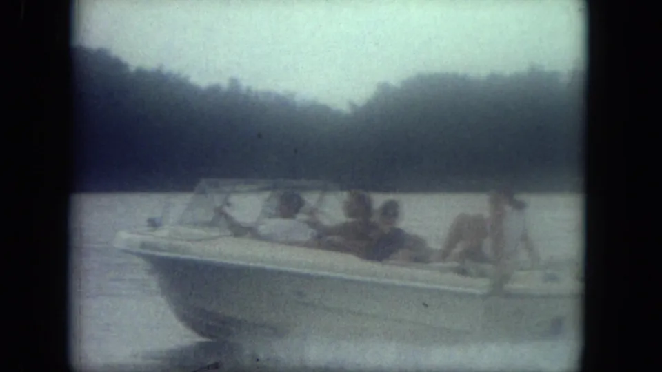 Mississippi 1971 8mm frame with several people riding in a white speedboat on calm water before a tree-lined shore.