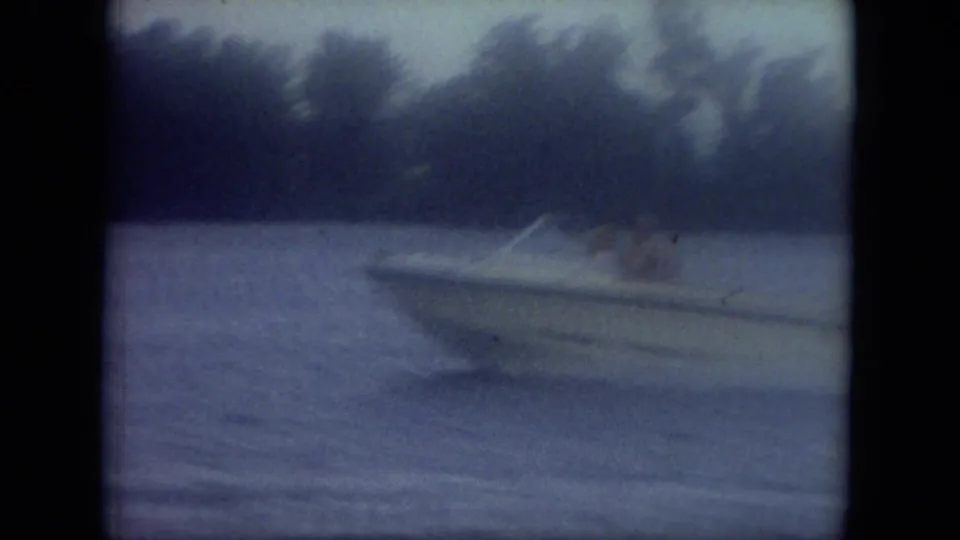 Mississippi 1971 home-movie frame showing a white speedboat moving across calm blue-gray water against a wooded shore.