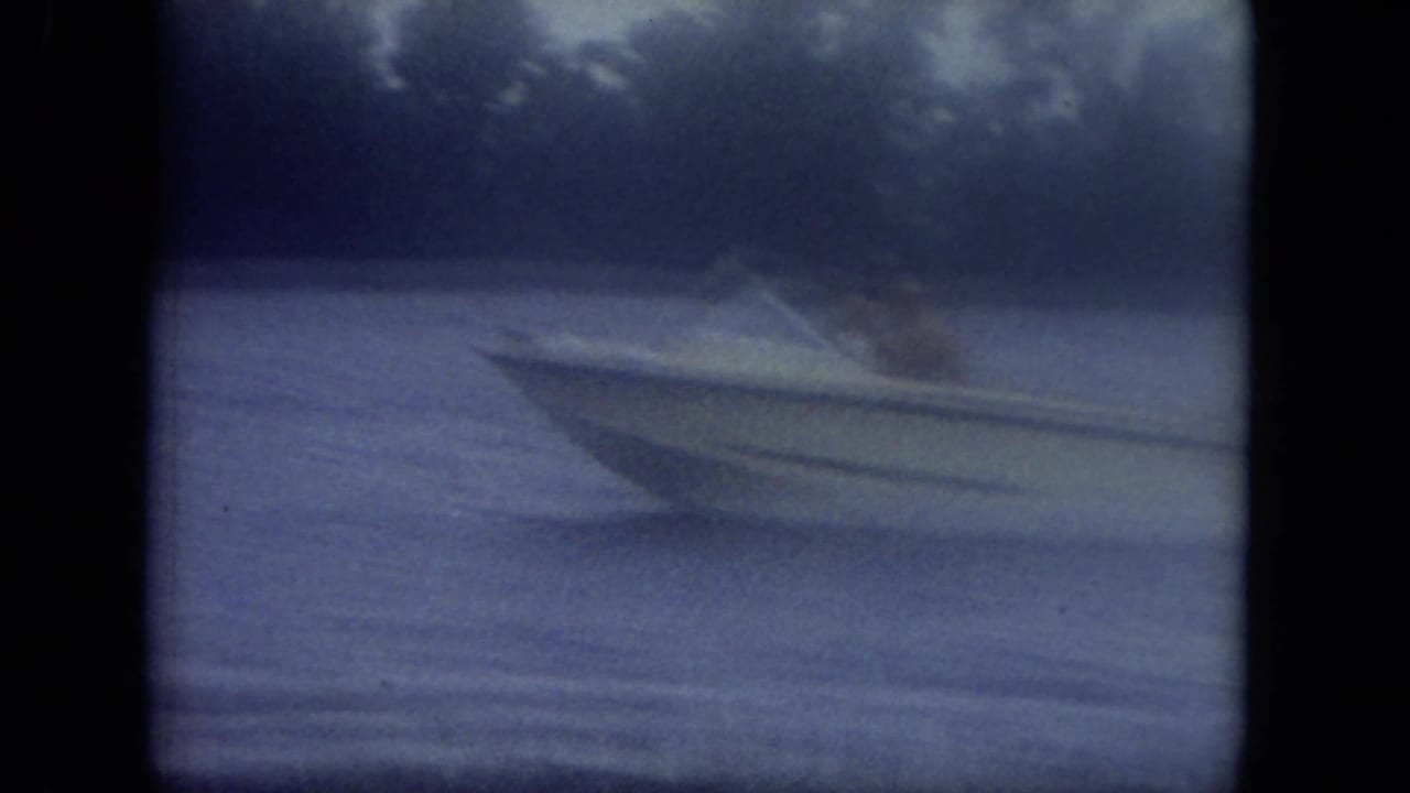 Speedboat moving across calm water near a dark tree-lined bank.