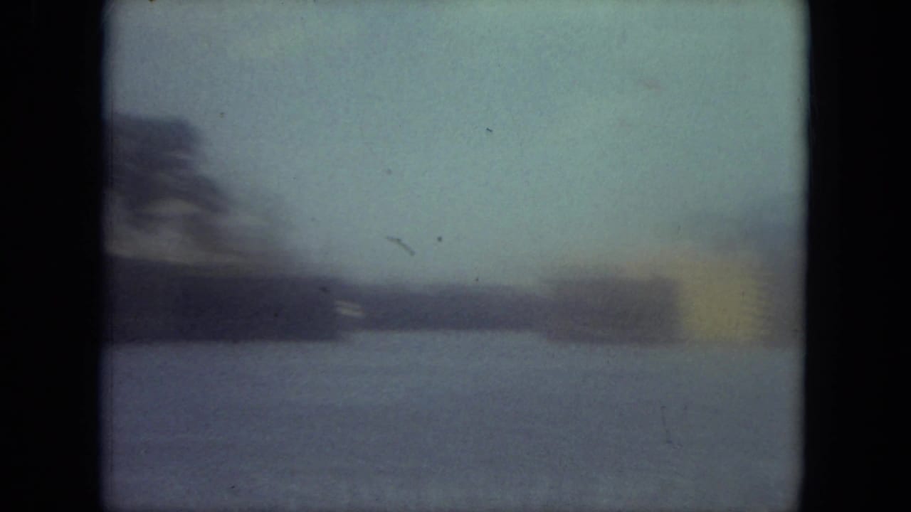 Soft blue-tinted 8mm frame of a Mississippi canal view with distant trees, low waterside structures, and open water.