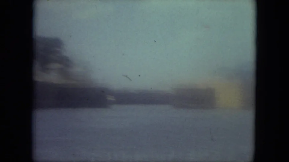 Soft blue-tinted 8mm frame of a Mississippi canal view with distant trees, low waterside structures, and open water.