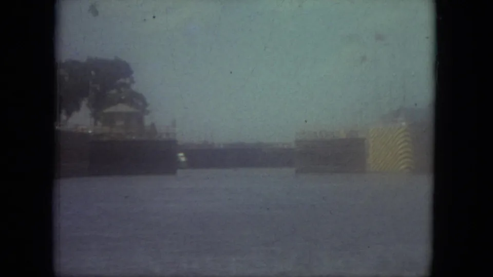 1971 Mississippi home-movie frame showing canal water, dark shoreline trees, low dock or launch structures, and a striped yellow object near the right edge.