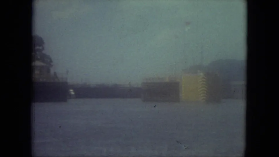 Blue-green 8mm frame from 1971 Mississippi showing a canal, distant waterside structures, shoreline trees, and a striped yellow object at right.