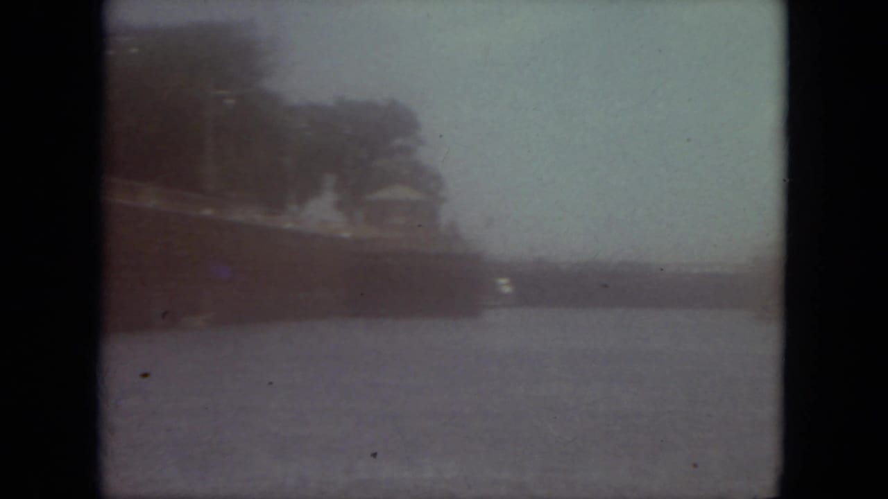 Hazy 8mm waterfront shot in Mississippi, with a distant shoreline and ship-like form across the water.