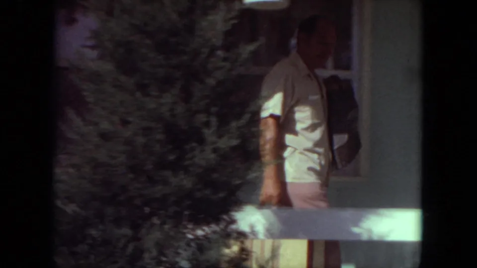 A man in a light short-sleeve shirt stands near a house porch, partly obscured by shrubs in 1971 home-movie footage.