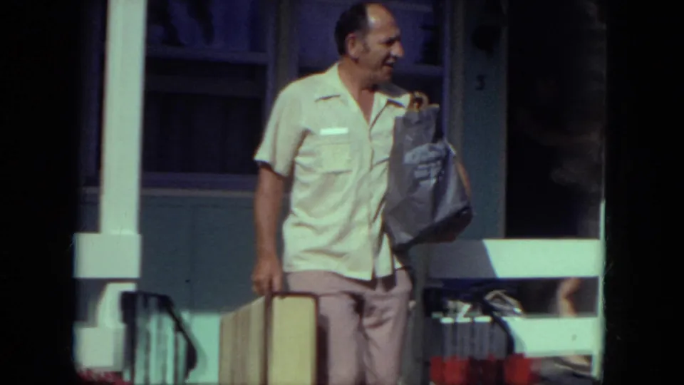 A man in a light shirt carries a suitcase and shoulder bag on a porch or front walk in 1971 Mississippi home-movie footage.