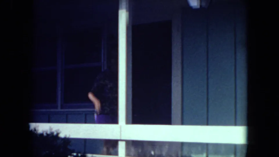 In Mississippi 1971 home-movie footage, a woman stands partly obscured on a porch beside windows, siding, and a white railing at night.