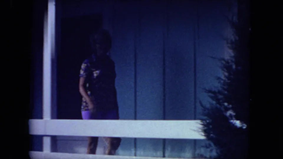 A woman in a patterned top and purple shorts stands on the porch of a blue house, framed by railing and dark foreground shadows.