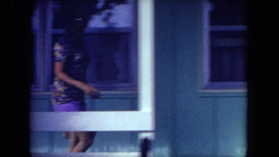 The woman passes between porch railings and windows on the blue house exterior, with strong shadow and 8mm color shift.
