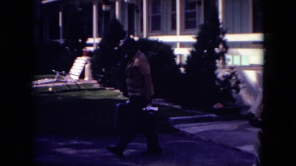 A man crosses the motel grounds in dim 8mm footage, moving past shrubs, lawn furniture, and the building exterior.
