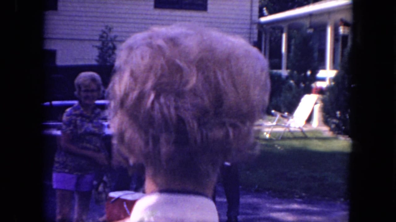 Mississippi 1971 home-movie frame showing adults outside a suburban house near a car, with a blonde woman viewed from behind.
