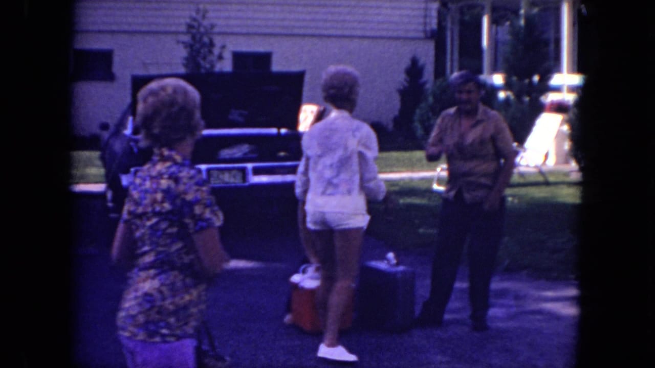 Mississippi 1971 frame of adults preparing luggage near a parked car with an open trunk outside a suburban house.