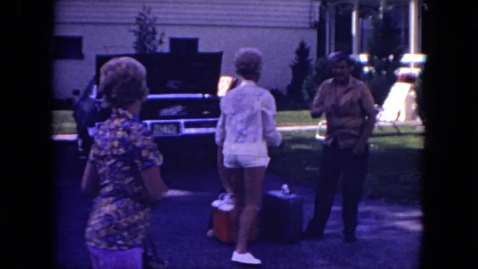 Mississippi 1971 frame of adults preparing luggage near a parked car with an open trunk outside a suburban house.