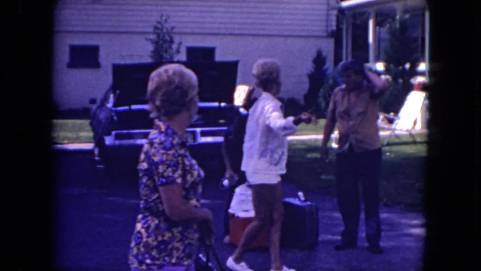 Mississippi 1971 home-movie frame with adults moving around luggage and a cooler near a car outside a house.