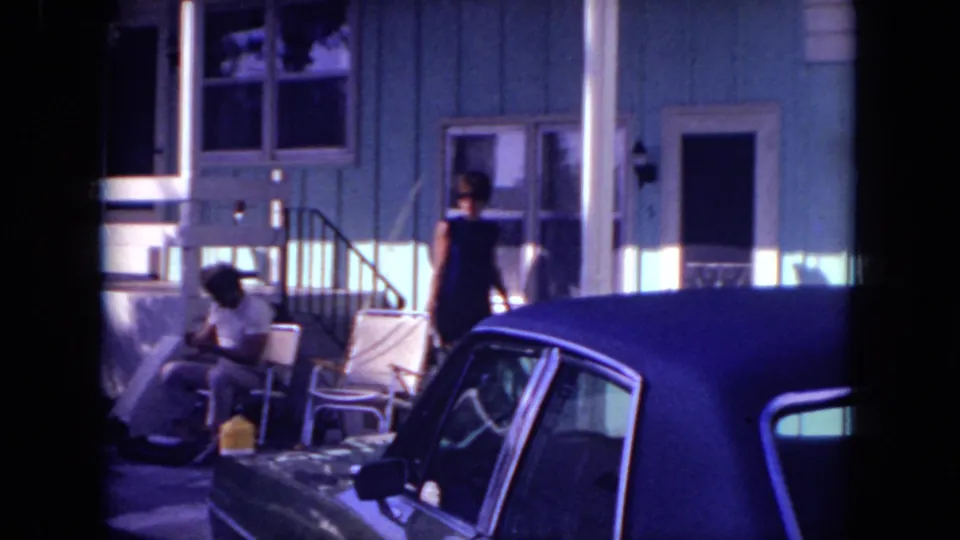 Mississippi 1971 frame showing a porch, lawn chairs, seated figure, standing woman, and parked car outside a house.