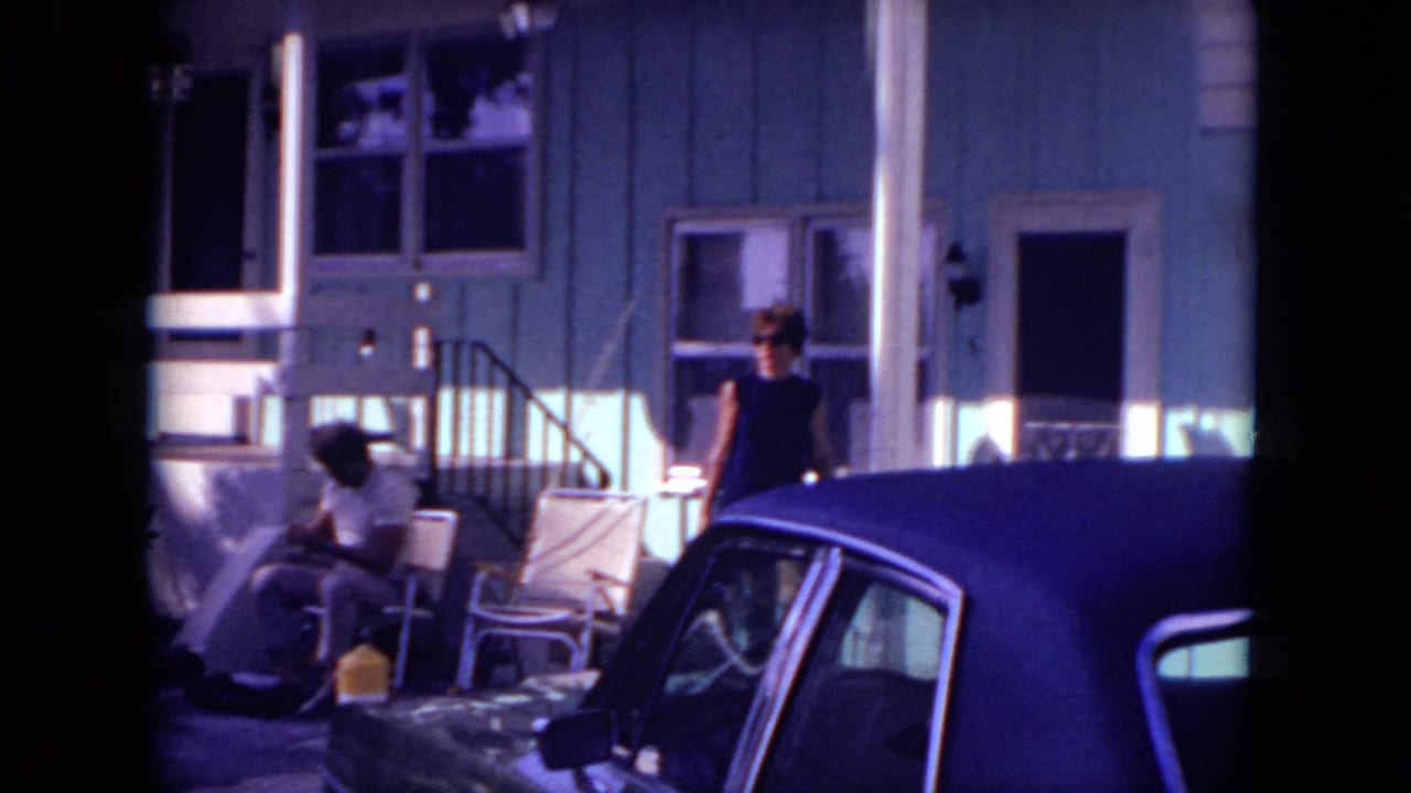 Woman in sunglasses stands near a dark-roofed sedan outside a house while another person sits on the porch.