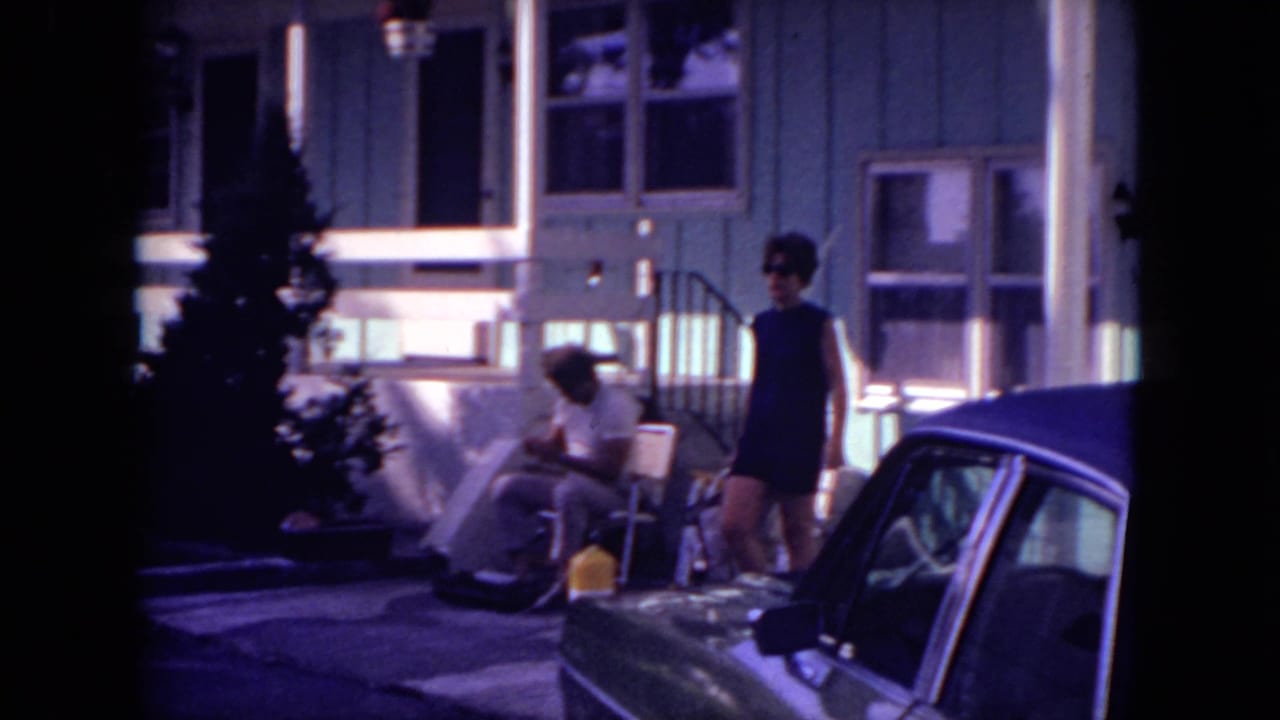 Woman in dark clothing stands by a parked car in front of a house with porch chairs and steps.